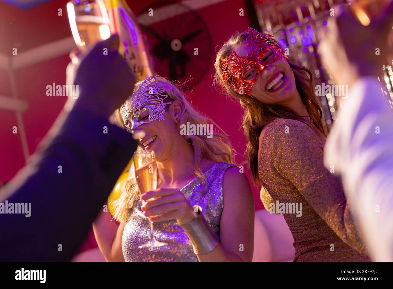 Two happy caucasian women wearing masks dancing and drinking champagne at a party in a nightclub. Fun, going out, celebrating and party concept. Stock Photo