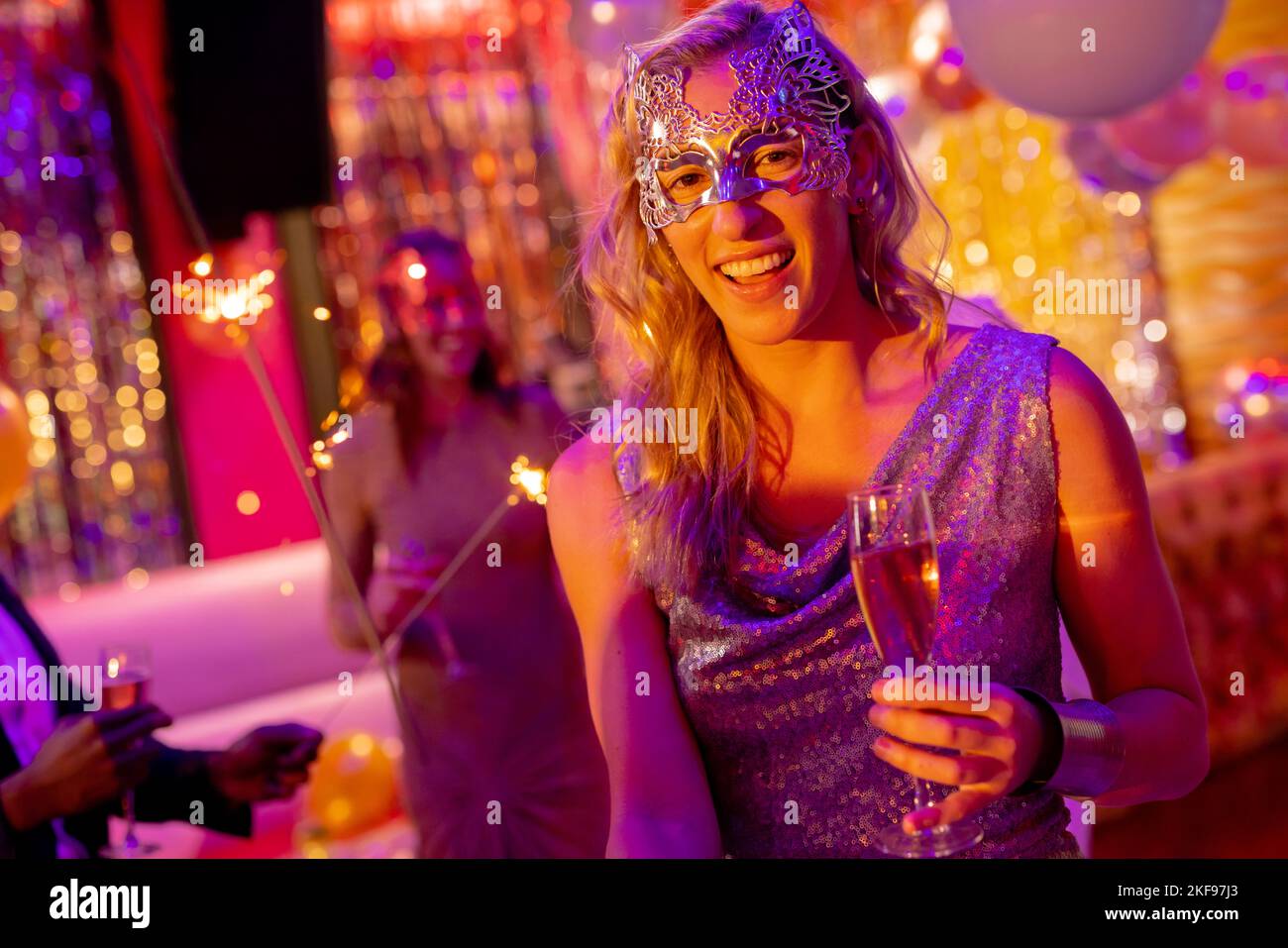 Portrait of smiling caucasian woman in mask holding glass of champagne at a party in a nightclub. Fun, going out, celebrating and party concept. Stock Photo