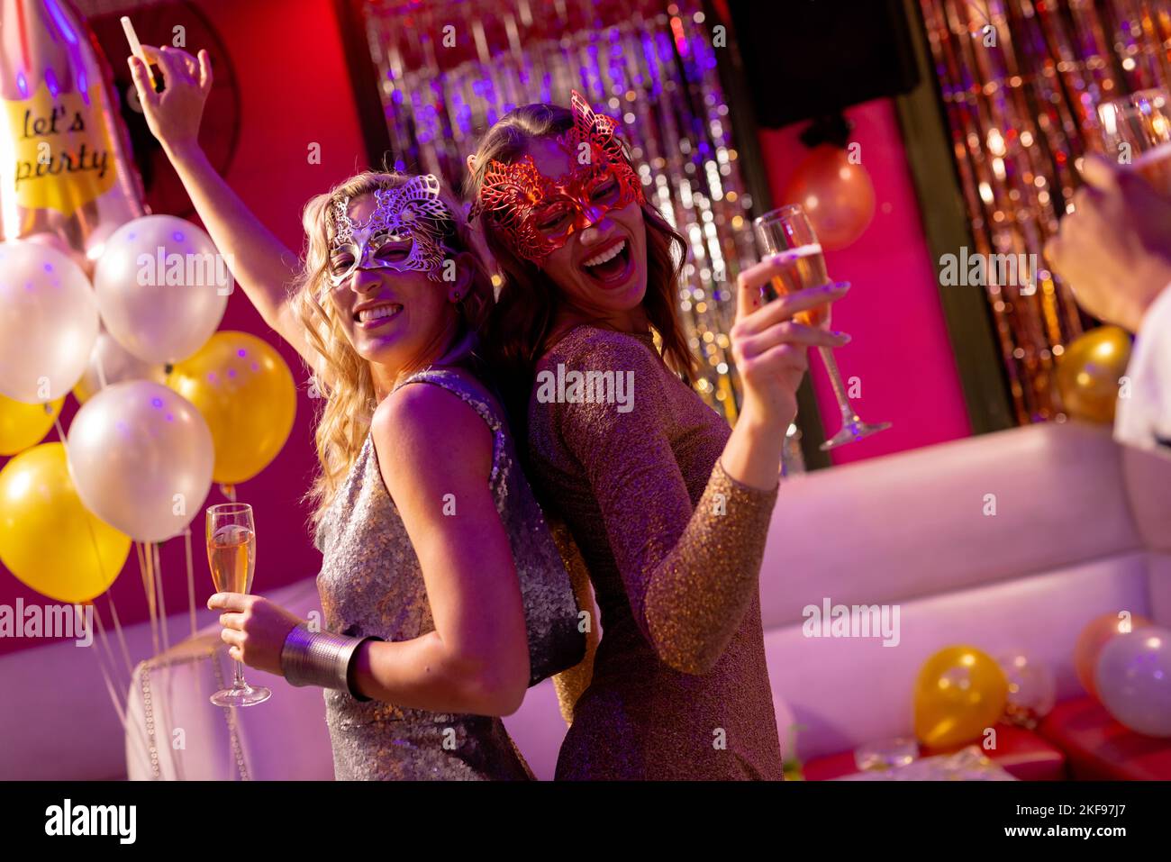 Two happy caucasian female friends in masks dancing and drinking champagne at a party in a nightclub. Fun, going out, celebrating and party concept. Stock Photo