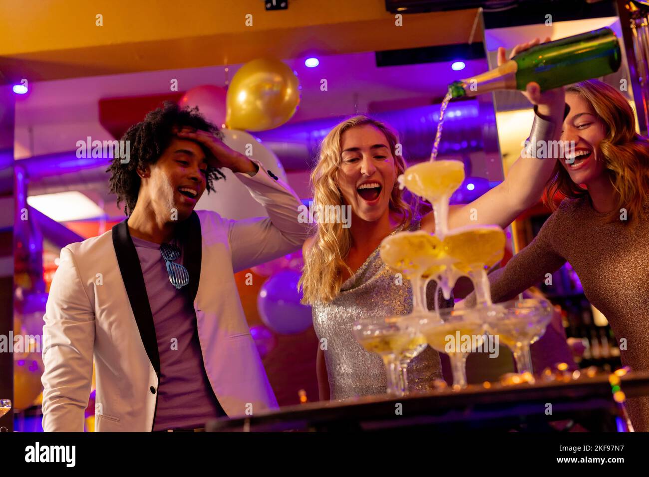 Happy caucasian woman pouring a champagne fountain into glasses with diverse friends at a nightclub. Fun, drinking, going out, inclusivity and party c Stock Photo