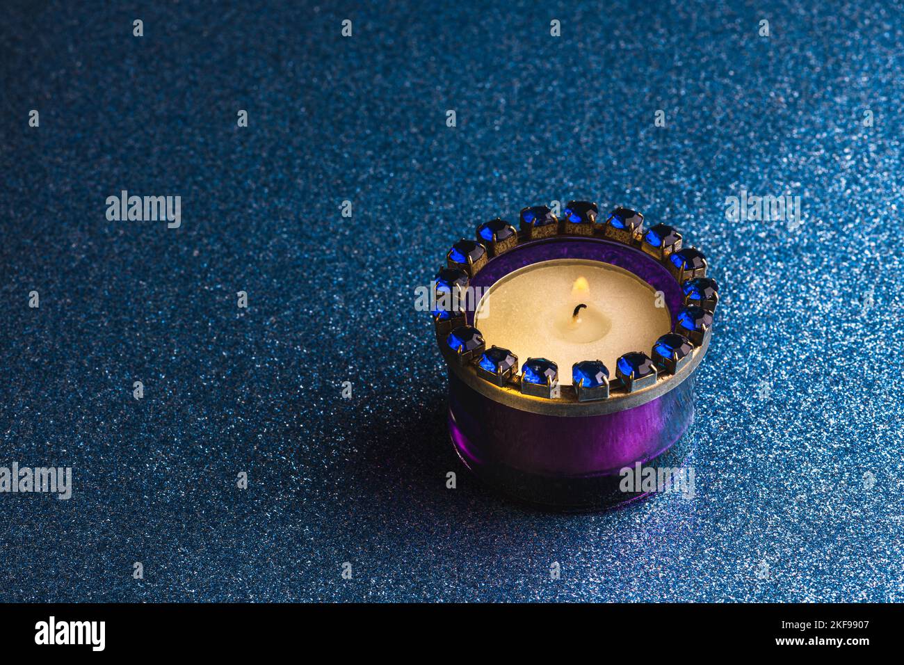 A burning tea light candle in a purple glass holder against a dark blue background with glitter. Stock Photo