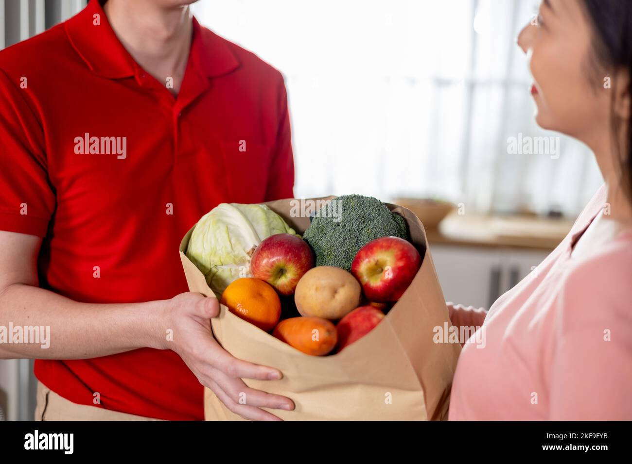 Asian delivery man in red uniform giving paper bag of food, fruit
