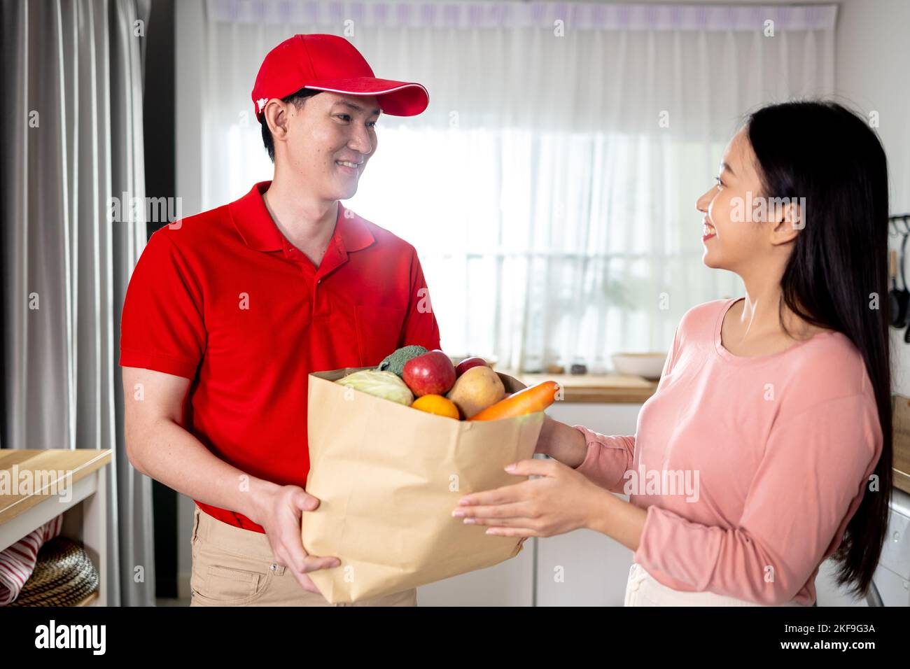 Asian delivery man in red uniform giving paper bag of food, fruit