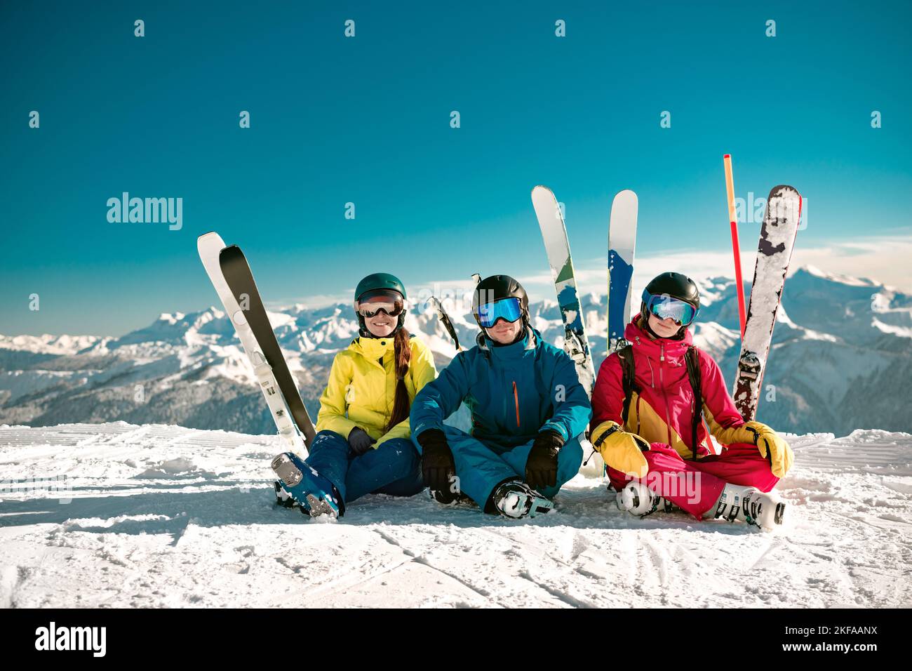 Three happy young skiers sits at mountain top and smiles. Winter vacations concept at ski resort Stock Photo