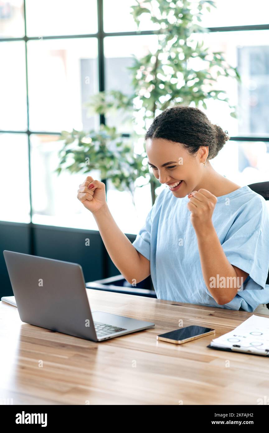 Vertical photo of joyful hispanic or brazilian business woman, sits at a work desk with laptop in office, rejoicing in success, big profit, celebrate deal, gesturing with fists, looks at laptop, smile Stock Photo
