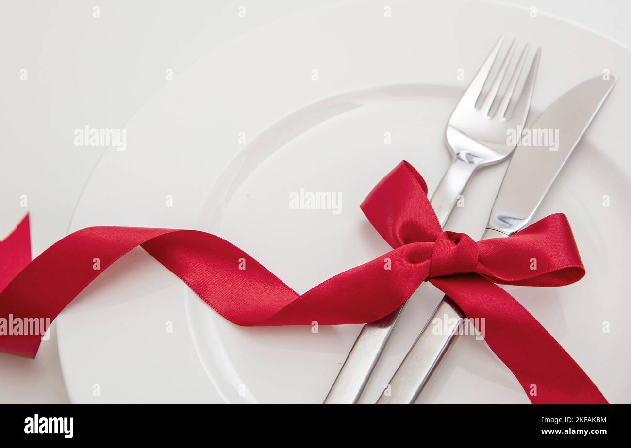 Christmas, Valentines day celebration dinner. Holiday table setting close up view. Cutlery and red satin ribbon decoration on white dishes, Stock Photo