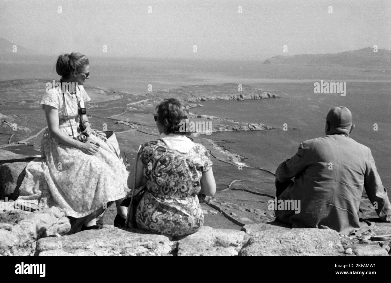 Griechenland - Ein Mann und zwei Frauen sitzen auf einer Klippe auf der Insel Delos, Kykladen, und blicken auf die Bucht, 1954. Greece - A man and two women sitting on a cliff at the island of Delos, Cyclades, have a look to the bay, 1954. Stock Photo