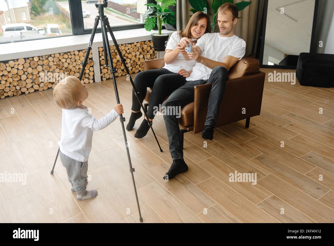 A child blogger holds a video tripod and prepares to become a young blogger. The parents are sitting next to each other in a chair. Stock Photo