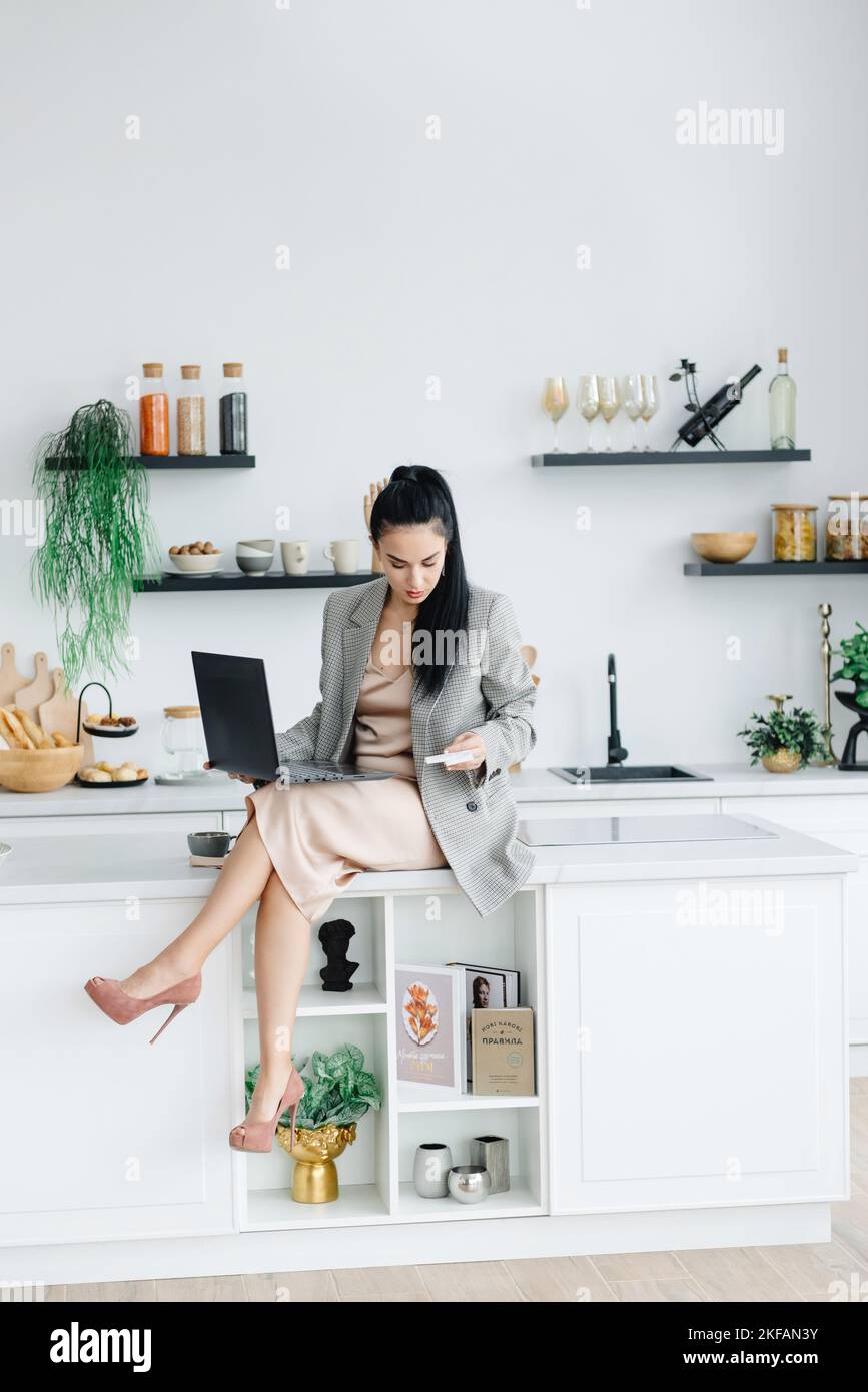 A young girl works from home. The woman in the kitchen works at her laptop to pay the bills. The stress of everyday life. Woman has internal health Stock Photo