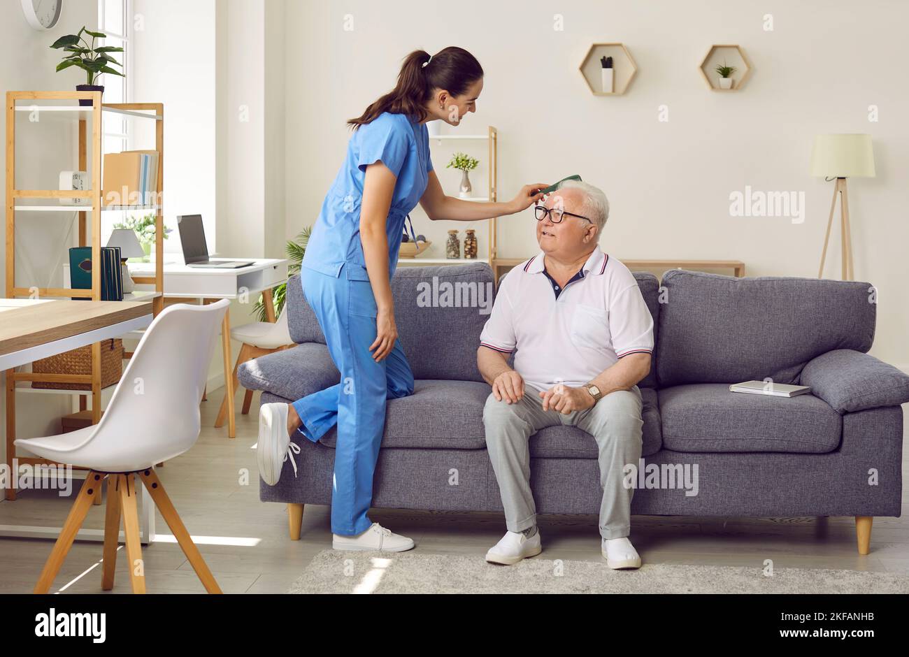 Friendly young female nurse combs hair of older man sitting on sofa in nursing home. Stock Photo