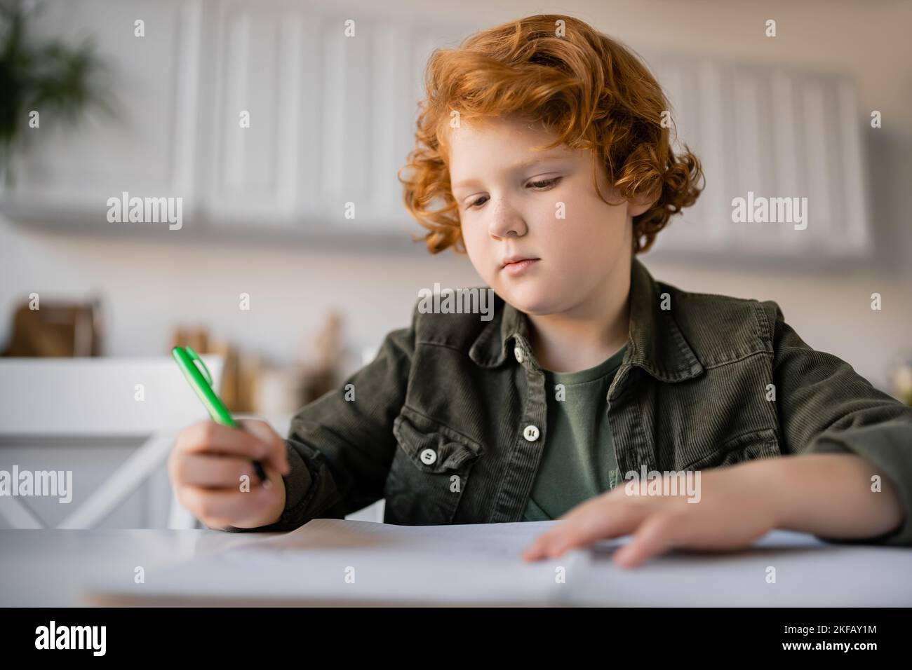 pensive redhead boy holding pen while doing homework near blurred notebook Stock Photo