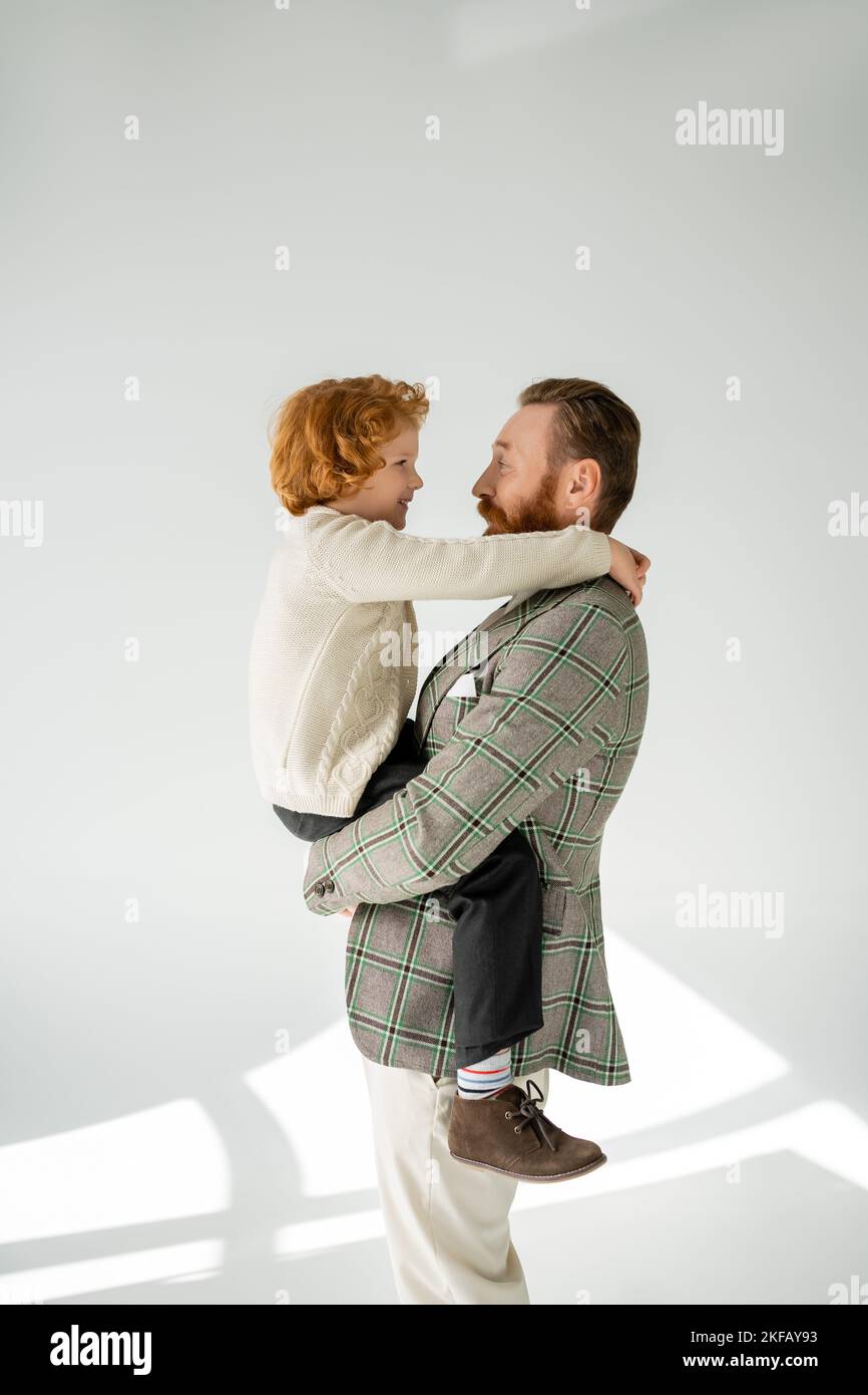 Side view of cheerful man in jacket hugging red haired son on grey ...