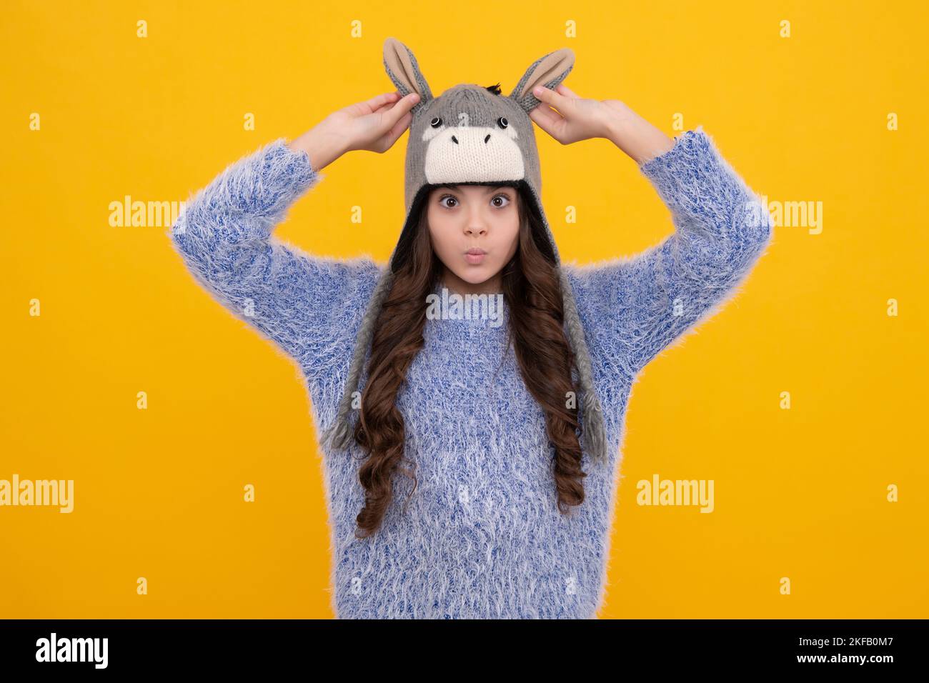 Beautiful winter kids portrait. Teenager girl posing with winter sweater and knit hat on yellow background. Funny face. Stock Photo