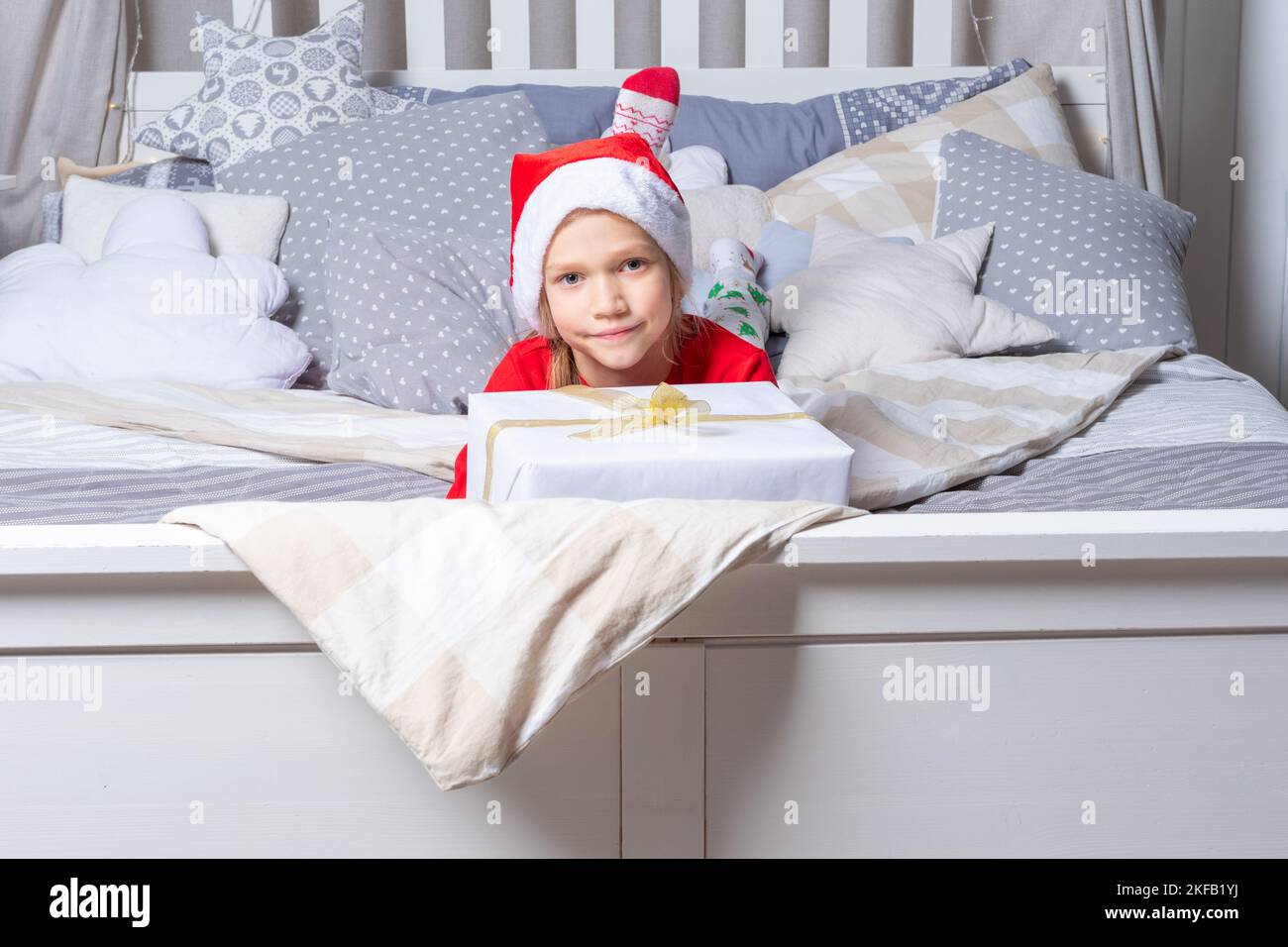 A girl in red pajamas and a Santa hat holds a large gift box, lying on the bed in the nursery. Christmas and New Year gifts for children. Family Chris Stock Photo