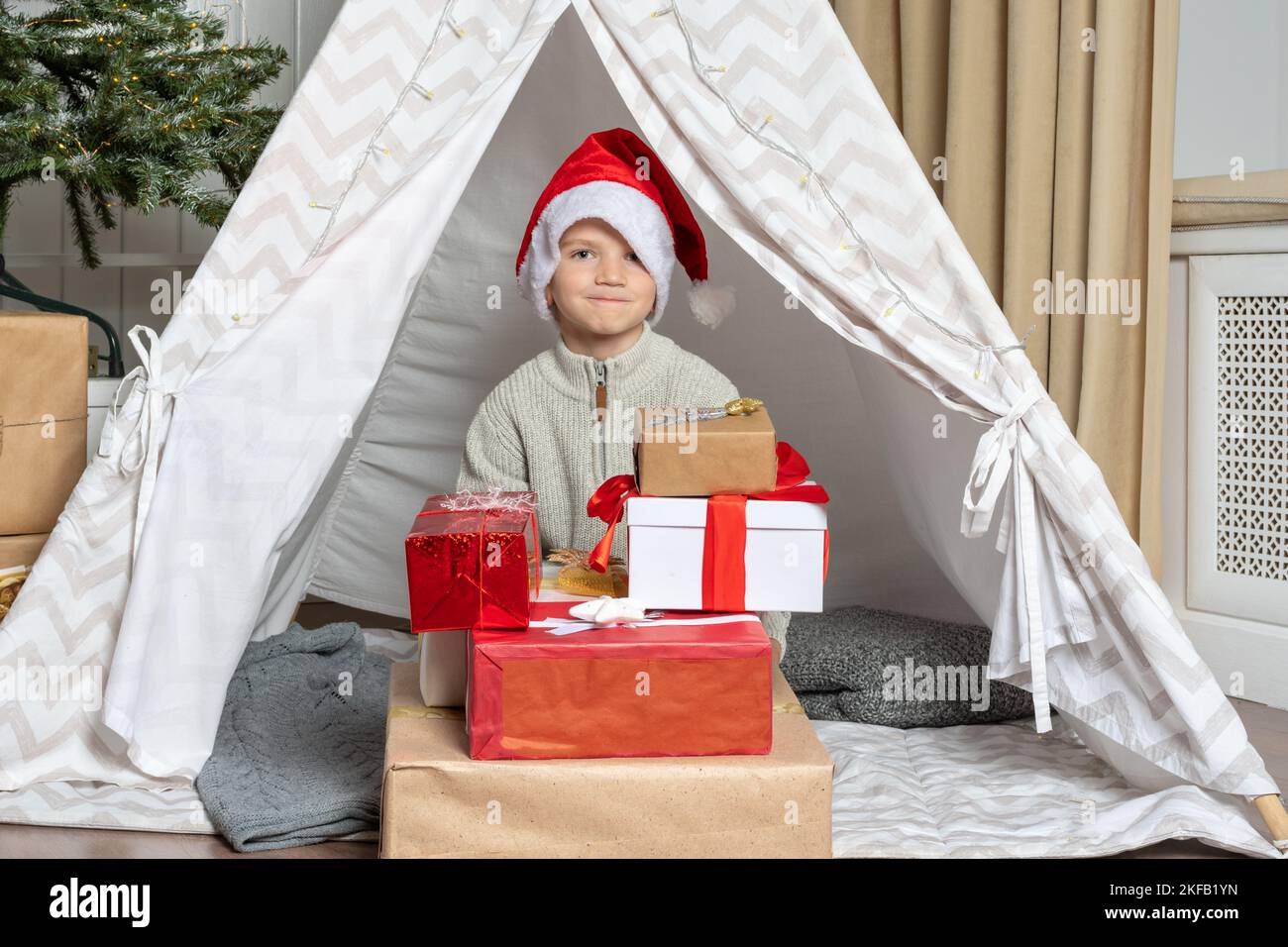A pretty preschooler boy in a Santa hat sitting on the floor next to a pile of his gifts in the nursery. The child is happy with gifts from Santa. Kid Stock Photo