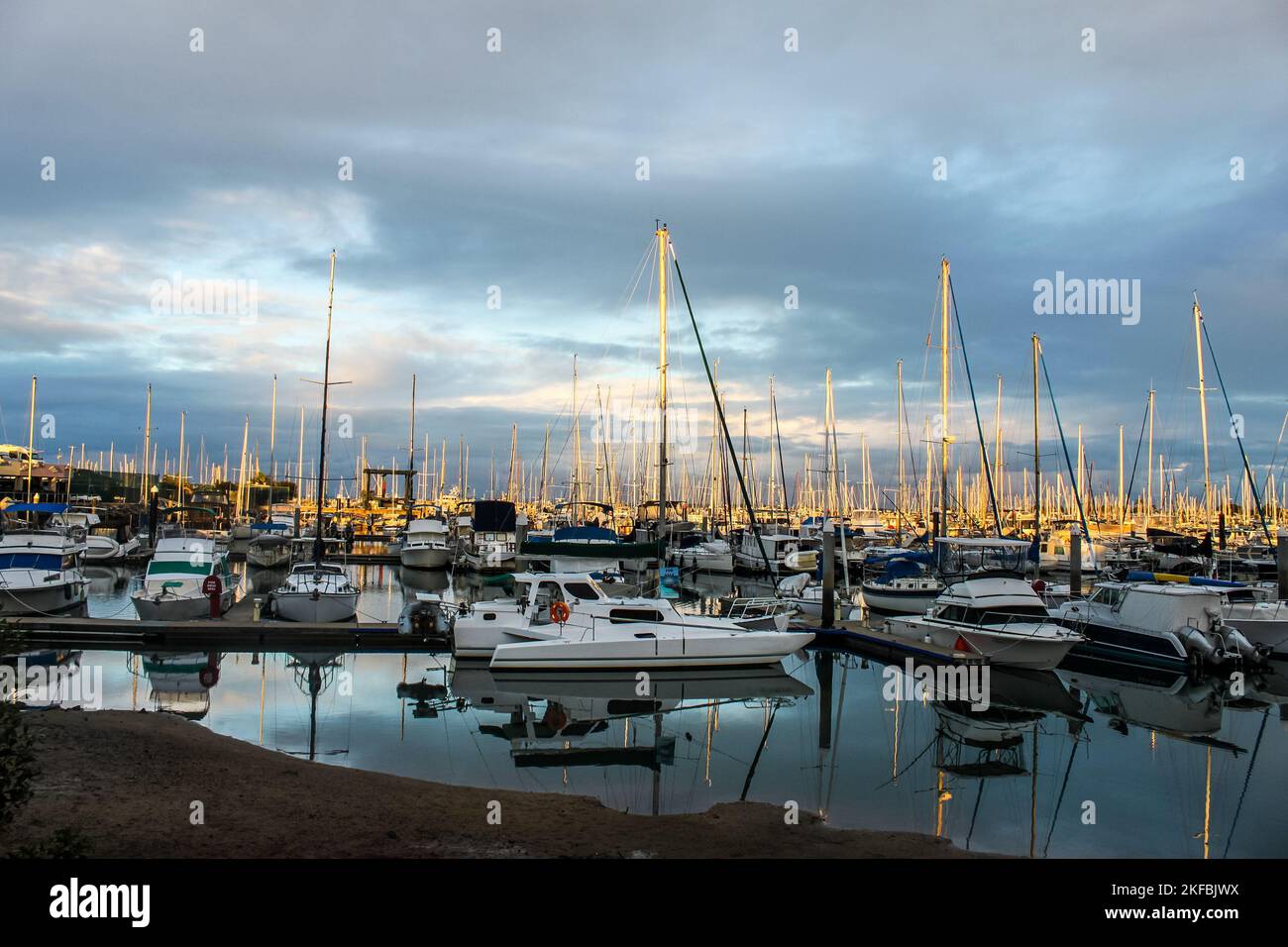 Many boats docked in marina at sunset with light shinning on masts and reflections in water. Stock Photo