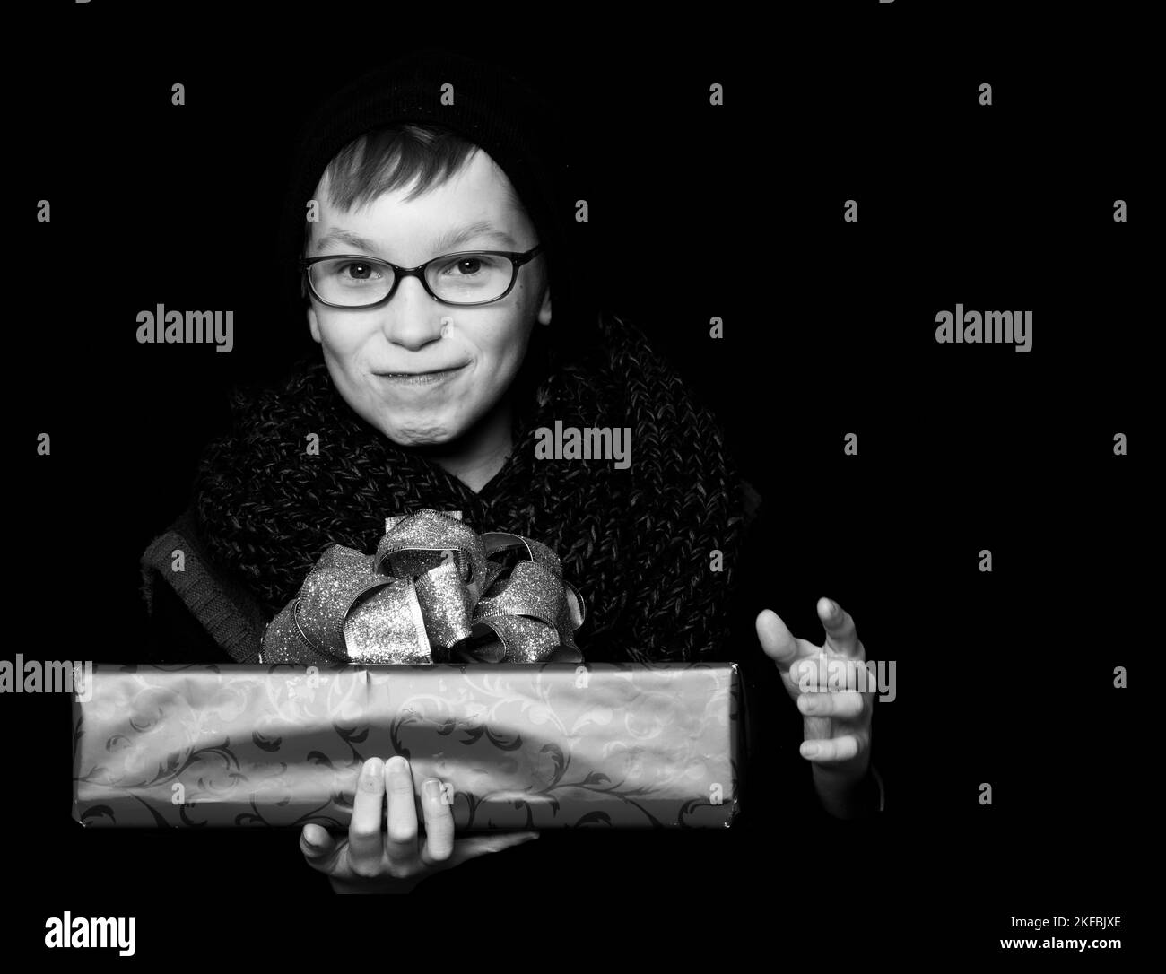 small smiling boy or cute nerd kid in glasses, hat and fashionable knitted scarf on black background holds red present box with golden bo Stock Photo