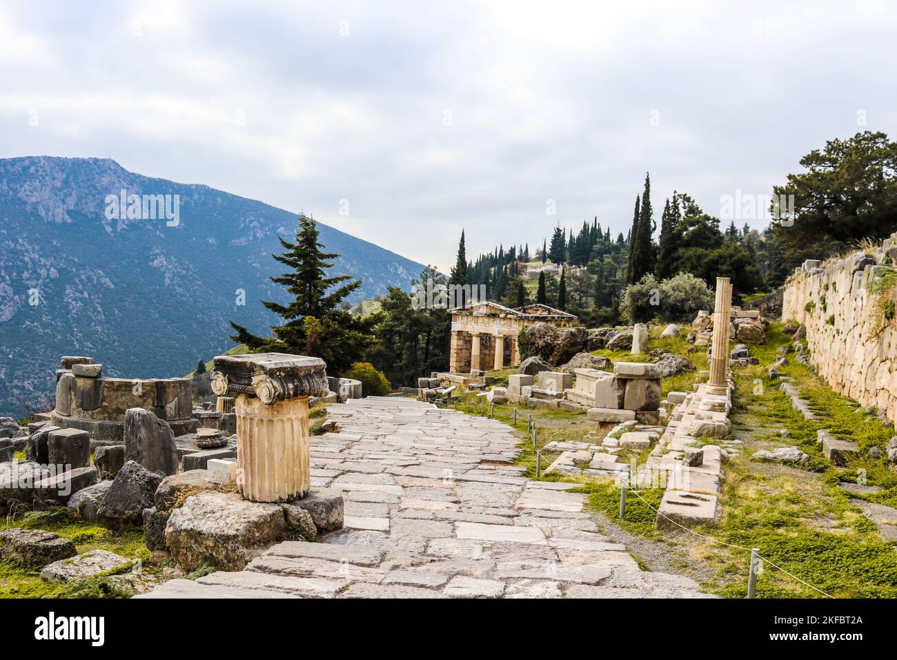 Rock walkway down hill in ancient Delphi Greece past ionic columns and parts of temples and past a reconstructed treasury with mountain in distance Stock Photo