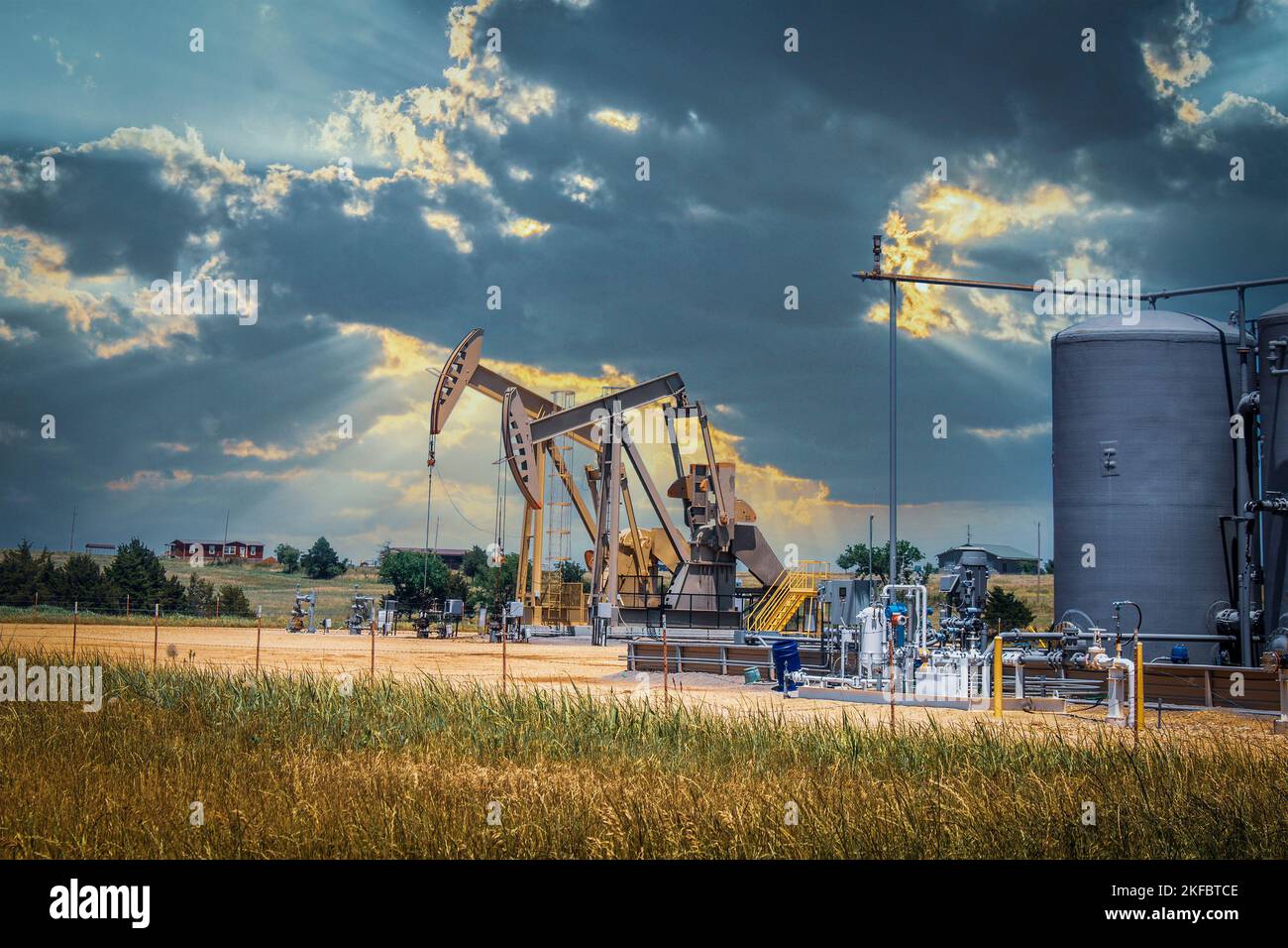 Two pump jacks and tanks and oil field equipment in fenced area in pasture under dramatic skies with houses on hill on horizon - summer Stock Photo