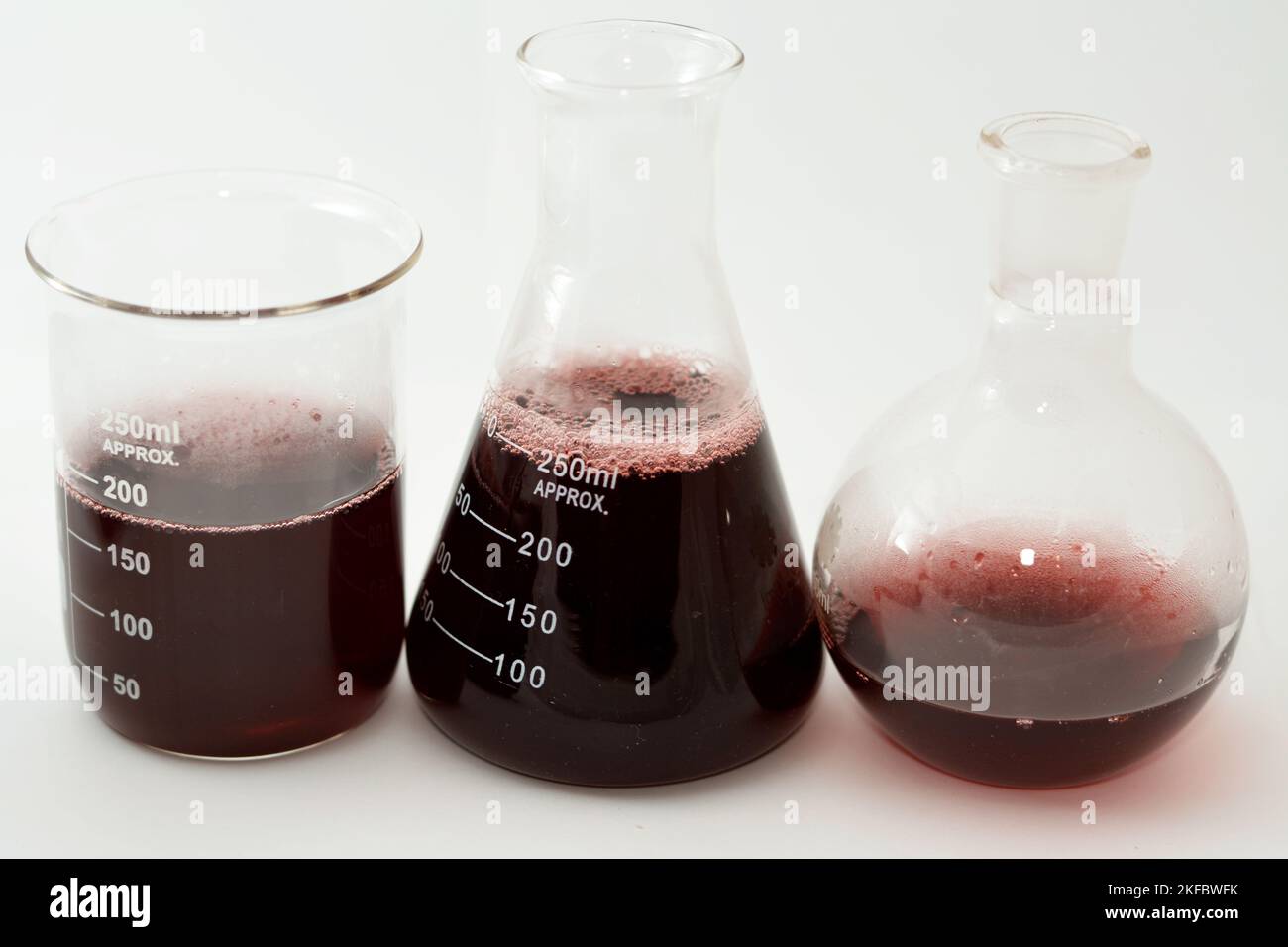 A laboratory set of glassware of conical, boiling and volumetric flasks that contains dark red liquid fluid with bubbles after a scientific experiment Stock Photo