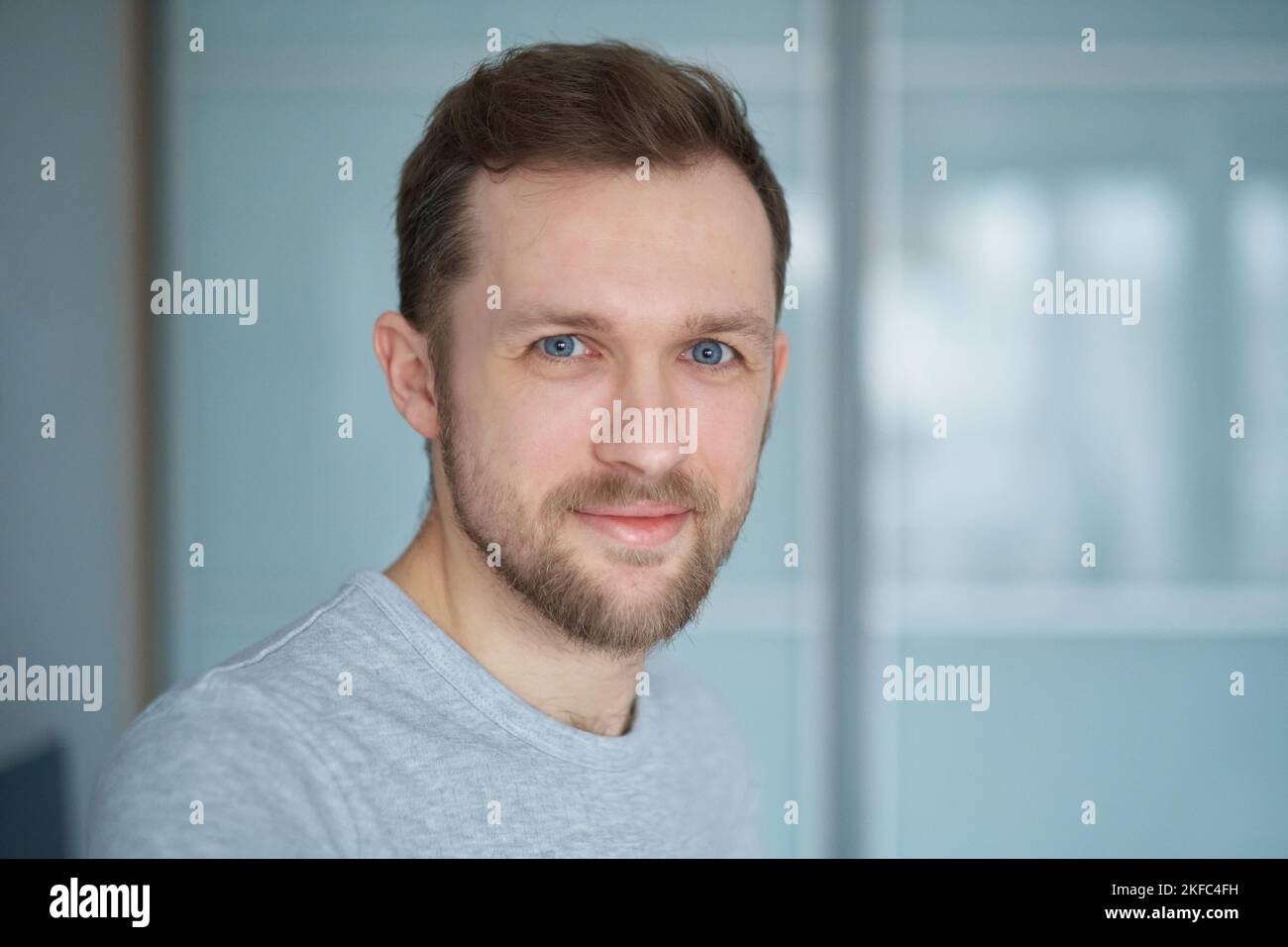 Close-up portrait of young handsome caucasian man in grey t-shirt looking in camera, positive emotion - smiling. Attractive bearded male in home office. Smart tech concept. High quality image Stock Photo