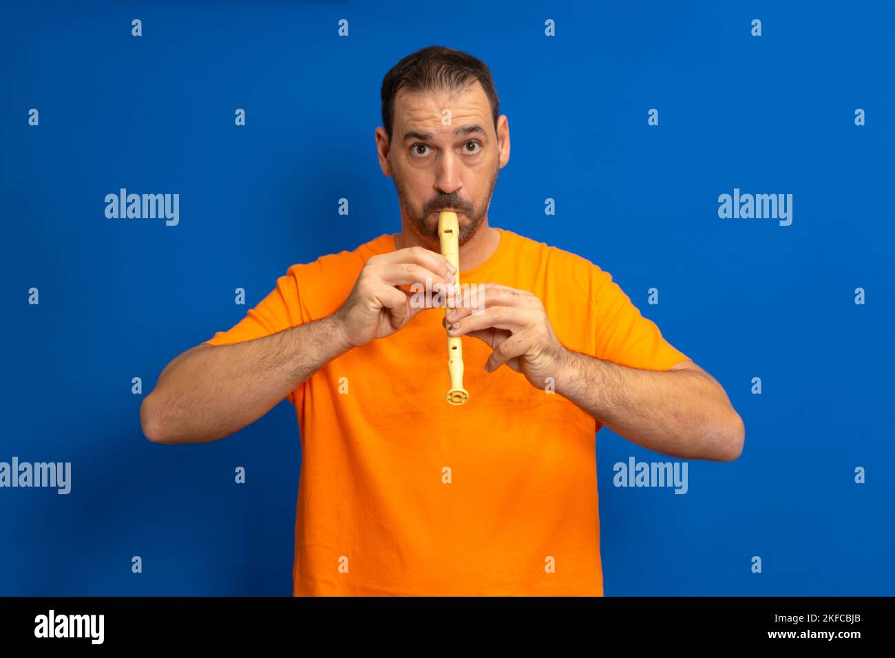 Hispanic man with a beard dressed in an orange t-shirt playing a school flute isolated over blue background Stock Photo