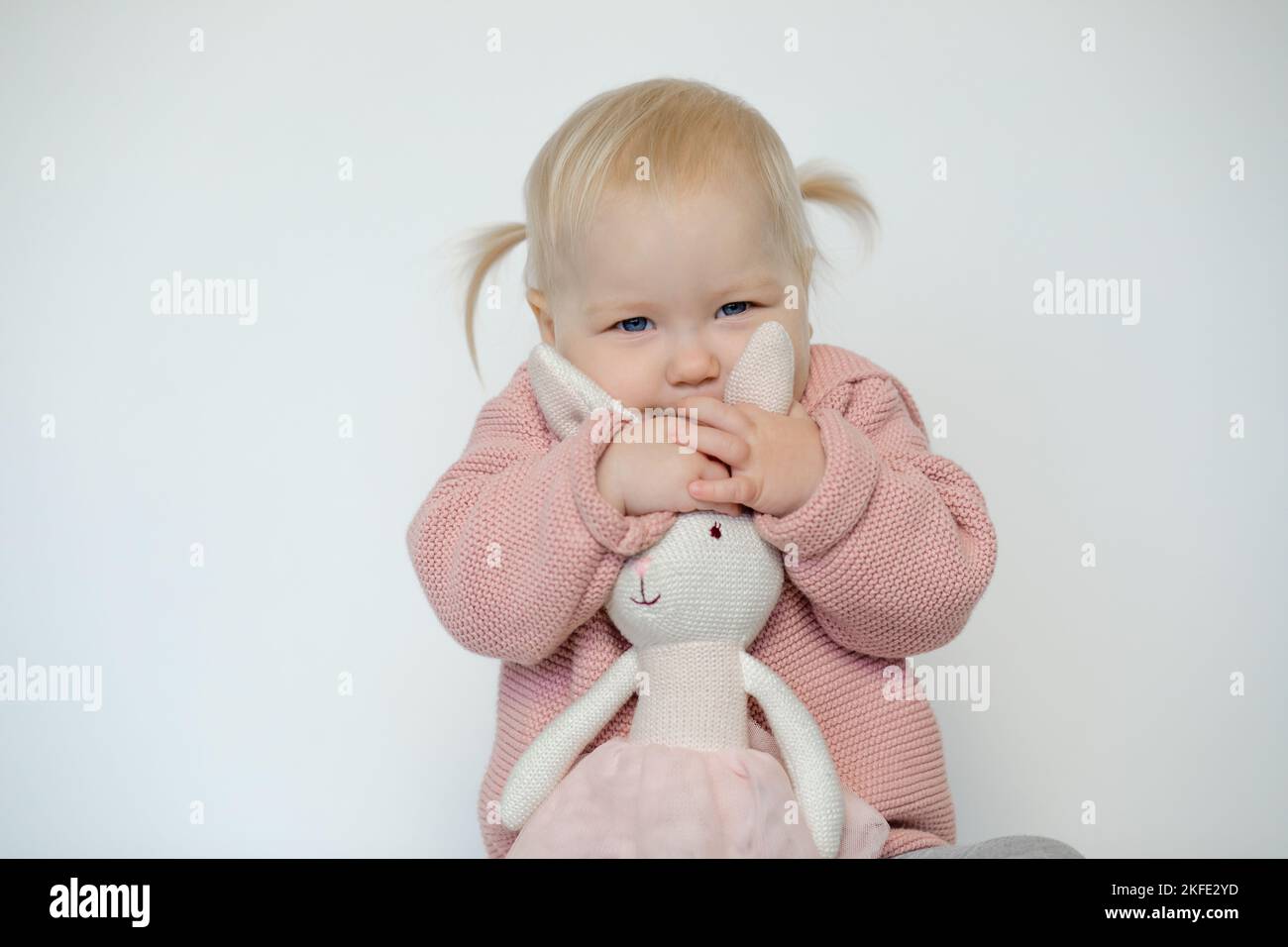 Pretty baby girl play with stuffed animal, isolated on white. Cheerful toddler tightly embracing teddy bunny. Blonde haired little child in pink Stock Photo