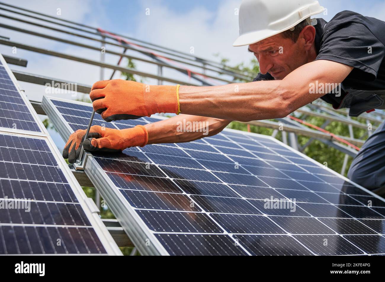 Close up of man installer placing solar module on metal rails. Male worker mounting photovoltaic solar panel system outdoors, wearing construction helmet and work gloves. Stock Photo