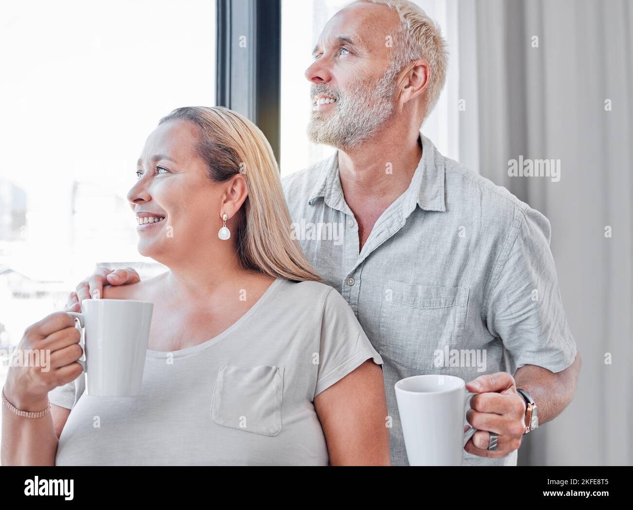 Elderly couple, hug and coffee by window with smile in contemplation, vision or morning routine at home. Happy senior man and woman smiling looking Stock Photo