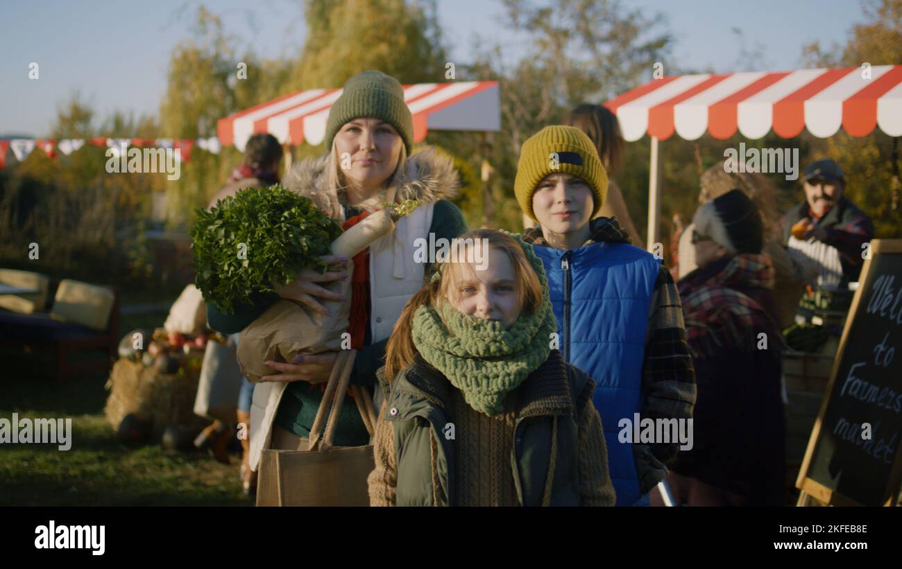 Mom with children's posing with bag of fruits or vegetables and looking at camera. Young girl and boy smiling and feeling happiness after shopping. Autumn fair outdoors. Stock Photo
