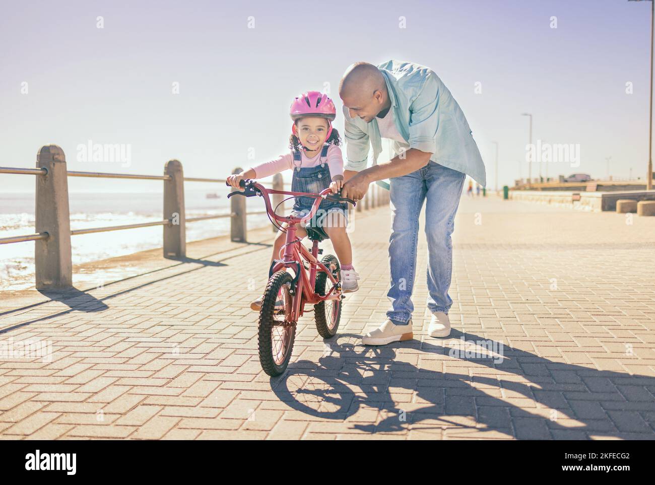 Father, child and bicycle with a girl learning to ride a bike on promenade by sea for fun, bonding and quality time on summer vacation. Man teaching Stock Photo