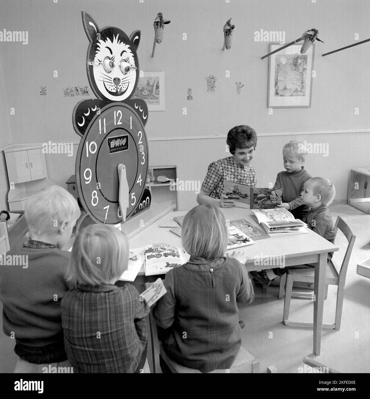 In the 1960s. The local Farsta Centrum shopping mall and area offers the parents shoppers a service of looking out for their children while doing the shopping. Pictured four children with a woman at a table reading and playing. A learning clock is visible, with extra large numbers for hours for the children to play and learn with.  Sweden 1967. Conard ref 5427 Stock Photo