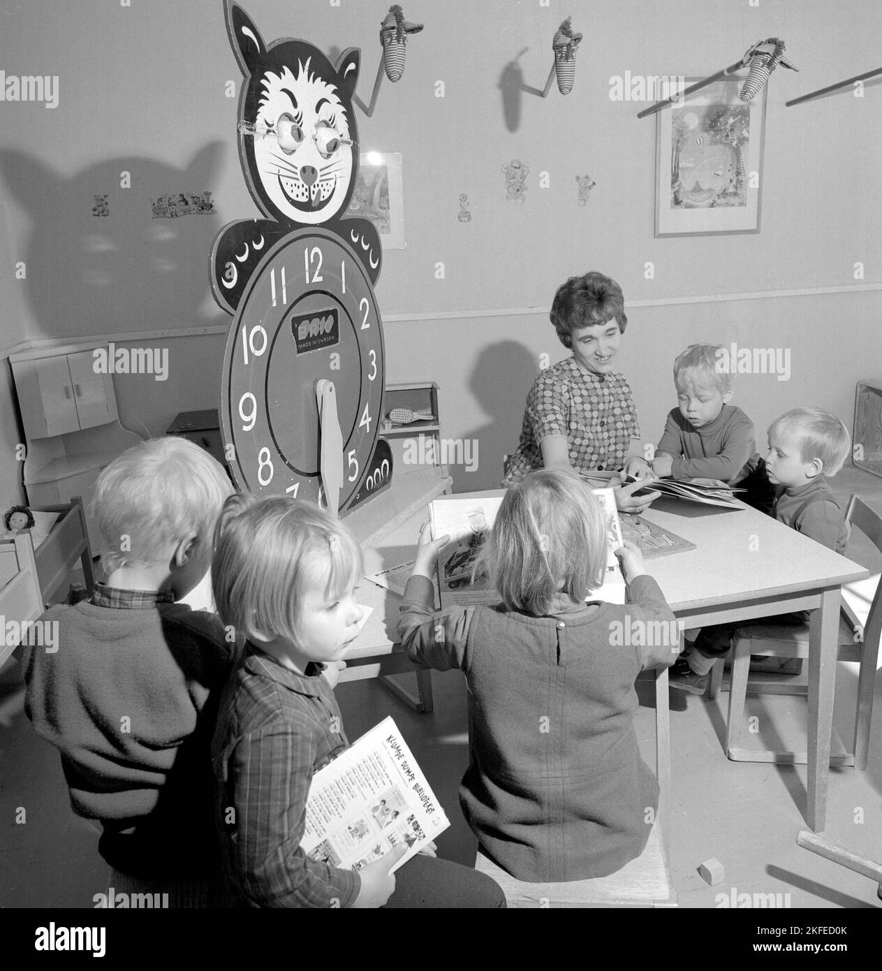 In the 1960s. The local Farsta Centrum shopping mall and area offers the parents shoppers a service of looking out for their children while doing the shopping. Pictured four children with a woman at a table reading and playing. A learning clock is visible, with extra large numbers for hours for the children to play and learn with.  Sweden 1967. Conard ref 5427 Stock Photo