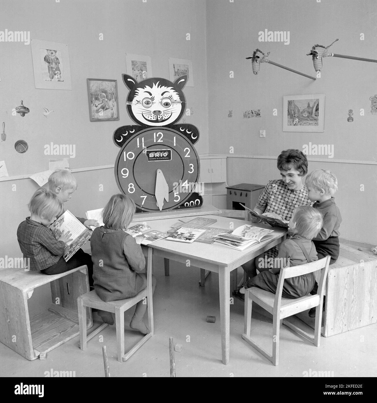 In the 1960s. The local Farsta Centrum shopping mall and area offers the parents shoppers a service of looking out for their children while doing the shopping. Pictured four children with a woman at a table reading and playing. A learning clock is visible, with extra large numbers for hours for the children to play and learn with.  Sweden 1967. Conard ref 5427 Stock Photo