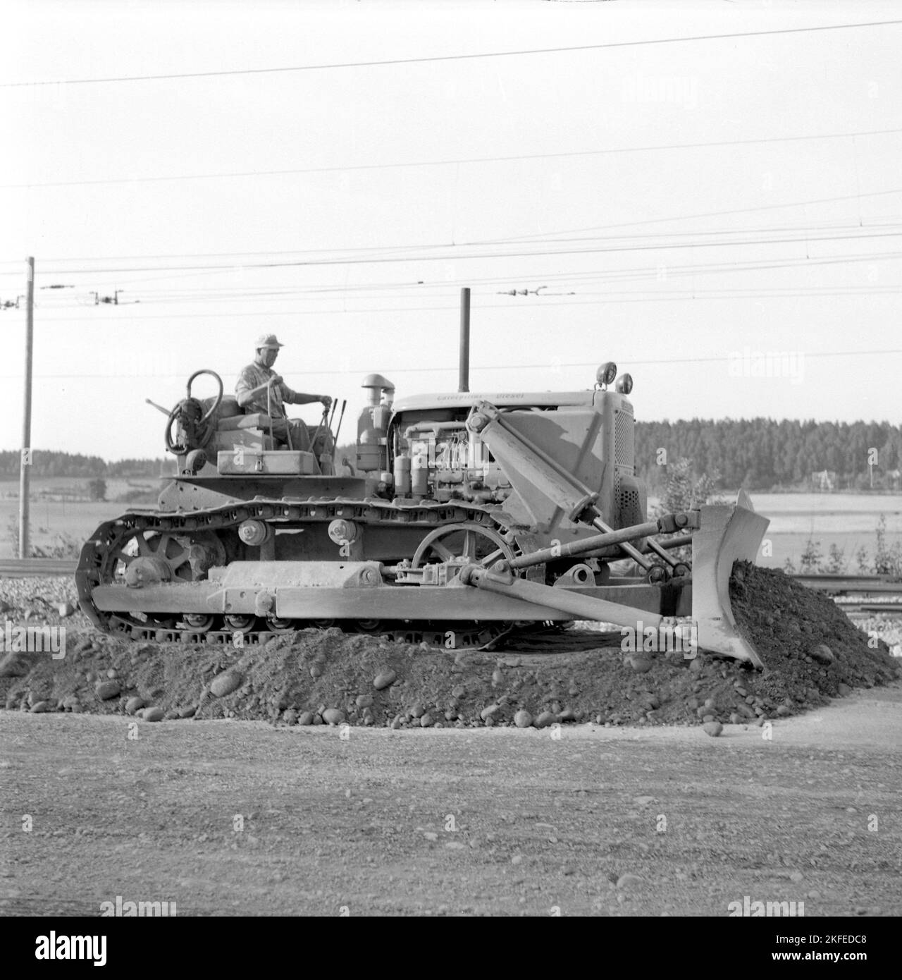 In the 1950s. The heavy machinery, a bulldozer looks gigantic when comparing to hte man sitting in the drivers seat with cabin or protection around him. It's a 20 tons Caterpillar Diesel model  D7, 240 hp engine. Cat is short for Caterpillar. Picture is taken on a railway construction site when the bulldozer flattens and spreads the material used to laying railway tracks on. Sweden august 1955 Stock Photo
