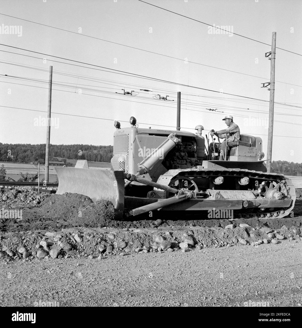 In the 1950s. The heavy machinery, a bulldozer looks gigantic when comparing to hte man sitting in the drivers seat with cabin or protection around him. It's a 20 tons Caterpillar Diesel model  D7, 240 hp engine. Cat is short for Caterpillar. Picture is taken on a railway construction site when the bulldozer flattens and spreads the material used to laying railway tracks on. Sweden august 1955 Stock Photo
