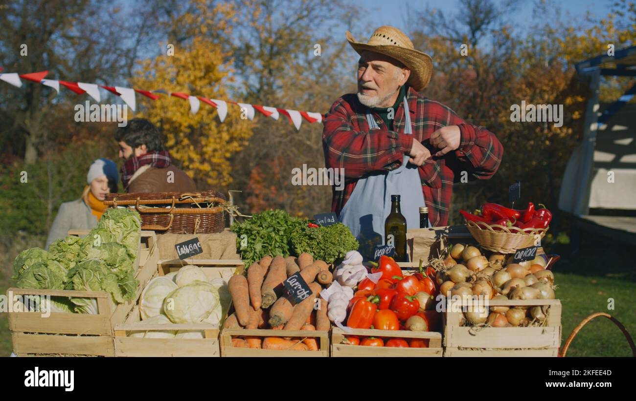 Senior farmer seller stands at the stall with fruits and vegetables, counts cash money. People shopping at local farmers market. Autumn fair. Organic food. Agriculture. Points of sale system. Stock Photo