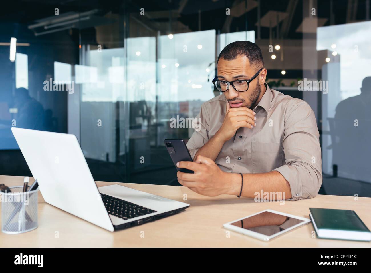 Thinking african american businessman reading news on smartphone, man working inside office at work using laptop, worker in shirt browsing online pages on phone serious and focused. Stock Photo