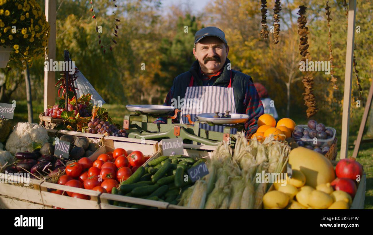 Elderly farmer stands at the stall with fresh colorful fruits and vegetables, looks at camera. Weekend shopping at local farmers market outside. Vegetarian, organic and healthy food. Agriculture. Stock Photo