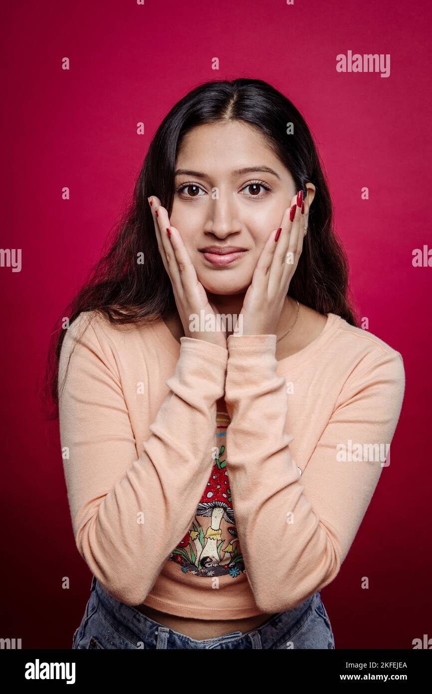 Portrait of happy Indian woman with hands on cheeks looking at camera against red background. Millennial girl with a beautiful and healthy skincare Stock Photo