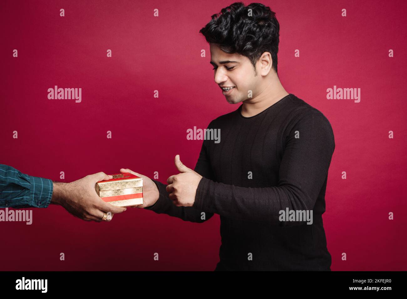 Happy Indian man in black t shirt gesturing thumbs up while receiving gift box from friend against red background Stock Photo