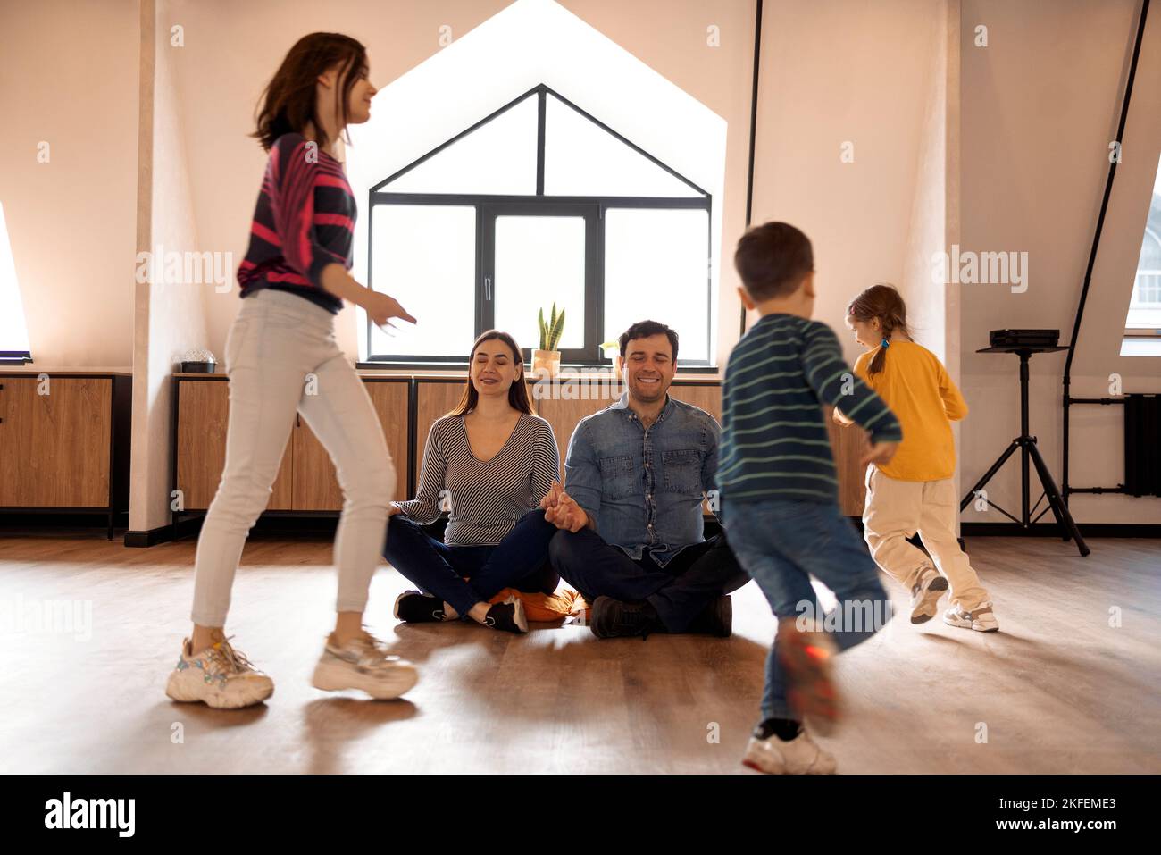Relaxing parents is sitting on the floor and meditating while there three children are running all over the apartment. While noisy daughters and son k Stock Photo