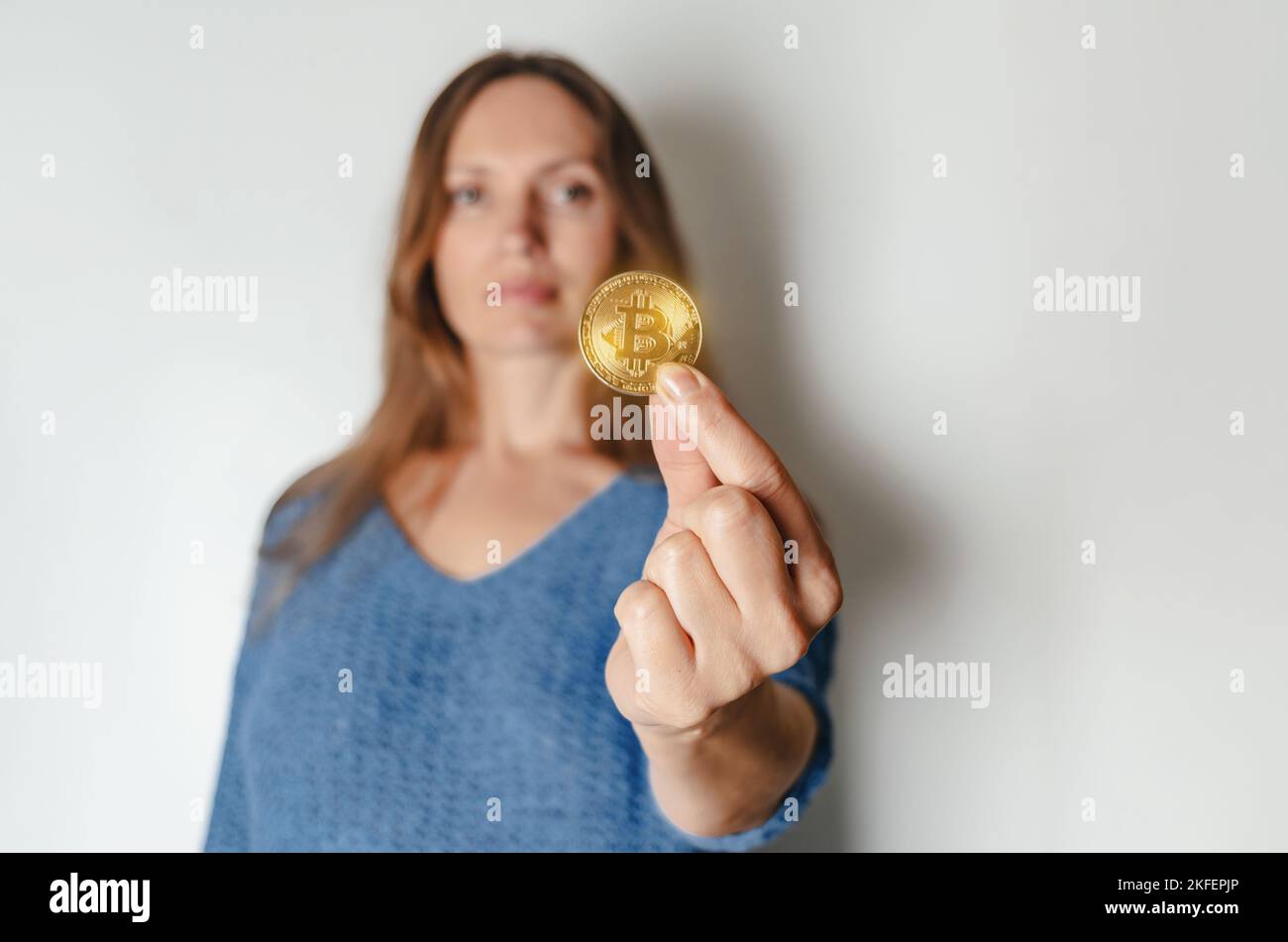 Joyful woman standing holding bitcoin paying attention to new digital cryptocurrency wearing blue sweater. Indoor studio shot isolated on white backgr Stock Photo