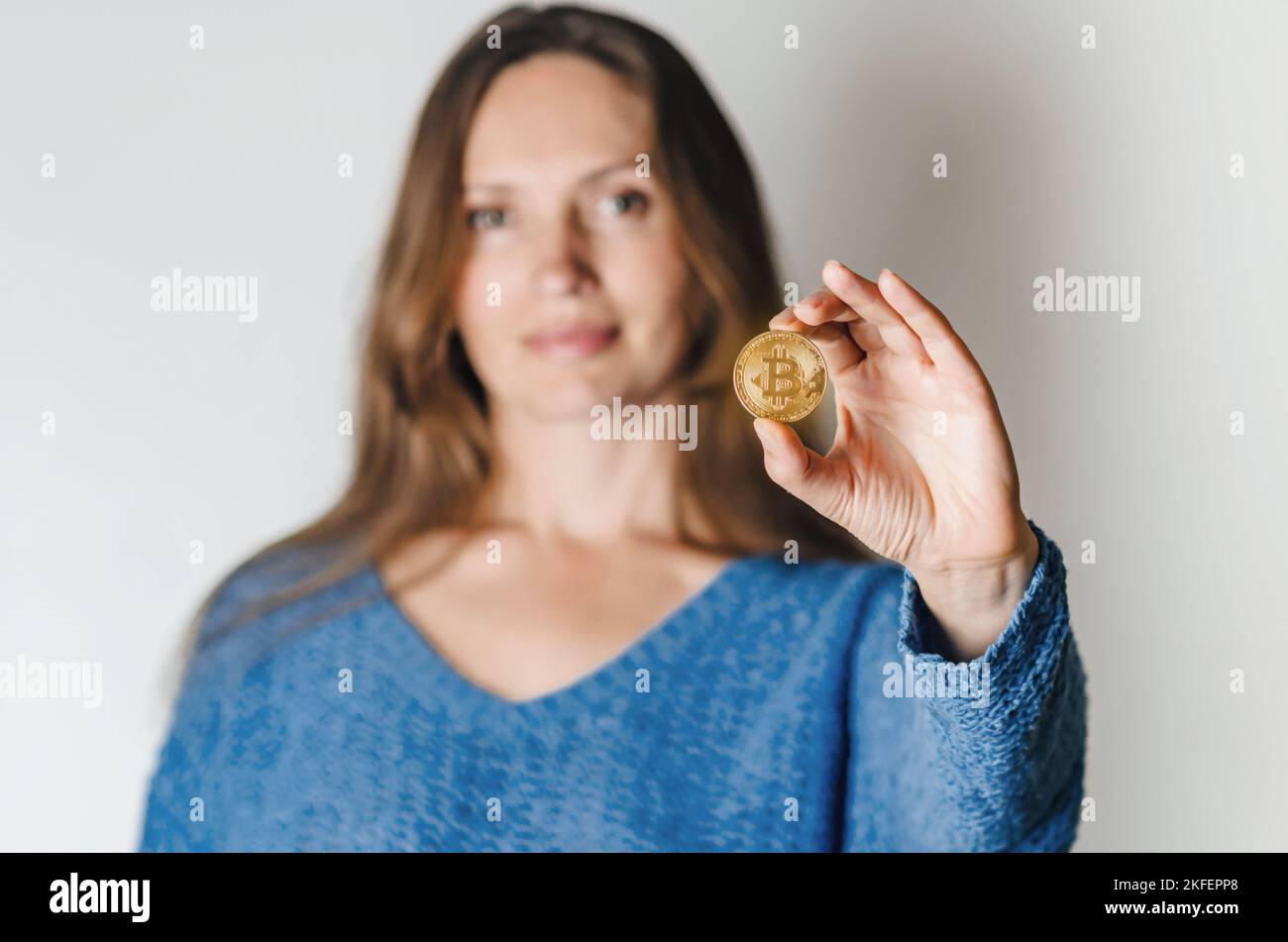 Joyful woman standing holding bitcoin paying attention to new digital cryptocurrency wearing blue sweater. Indoor studio shot isolated on white backgr Stock Photo