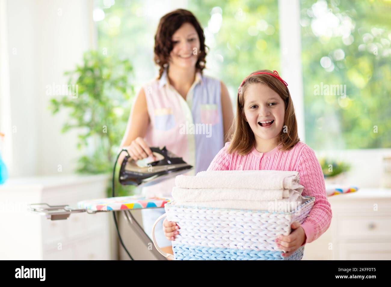 Woman and child ironing clothes. Mother and daughter folding clothes at iron board. Mom and kid cleaning house. Home chores. Stock Photo
