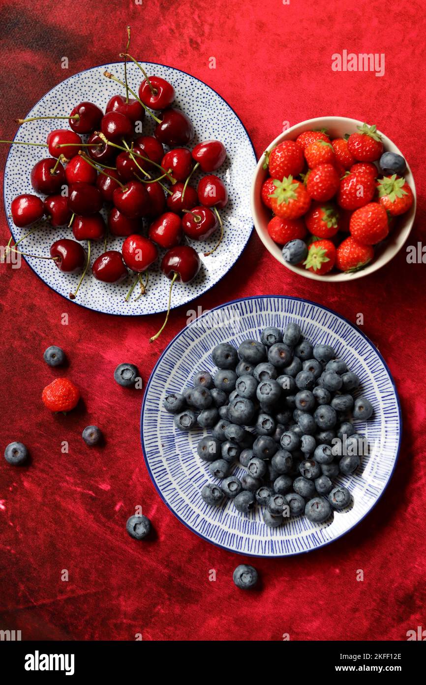 Vibrant colors of fresh summer berries. Strawberry, blueberry and sweet cherry on a blue ceramic plate. Red textured background with copy space. Stock Photo