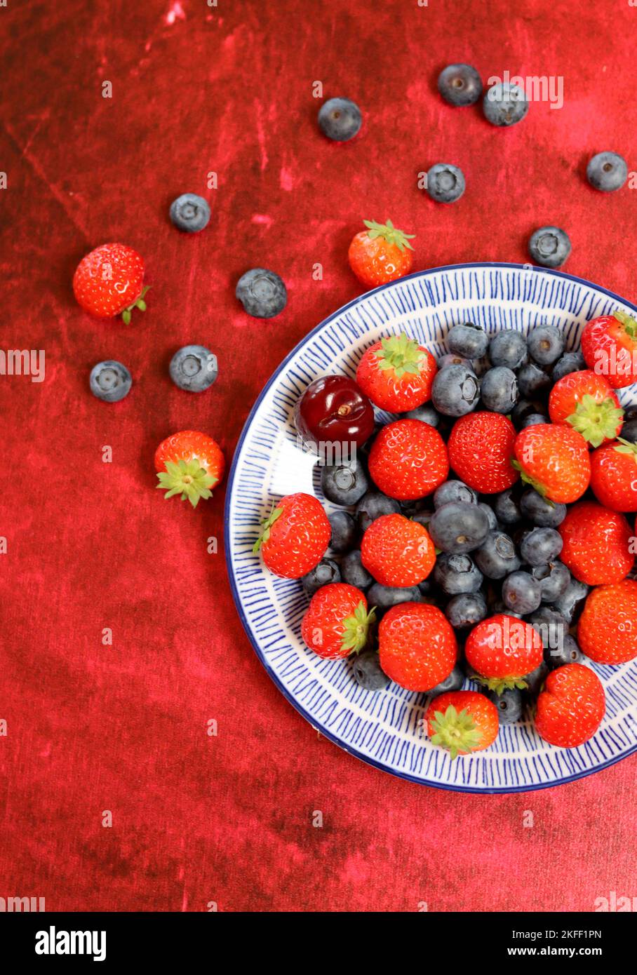 Vibrant colors of fresh summer berries. Strawberry, blueberry and sweet cherry on a blue ceramic plate. Red textured background with copy space. Stock Photo