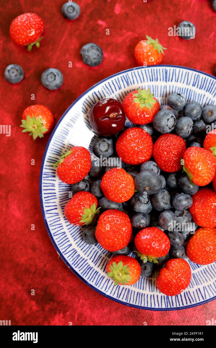 Vibrant colors of fresh summer berries. Strawberry, blueberry and sweet cherry on a blue ceramic plate. Red textured background with copy space. Stock Photo