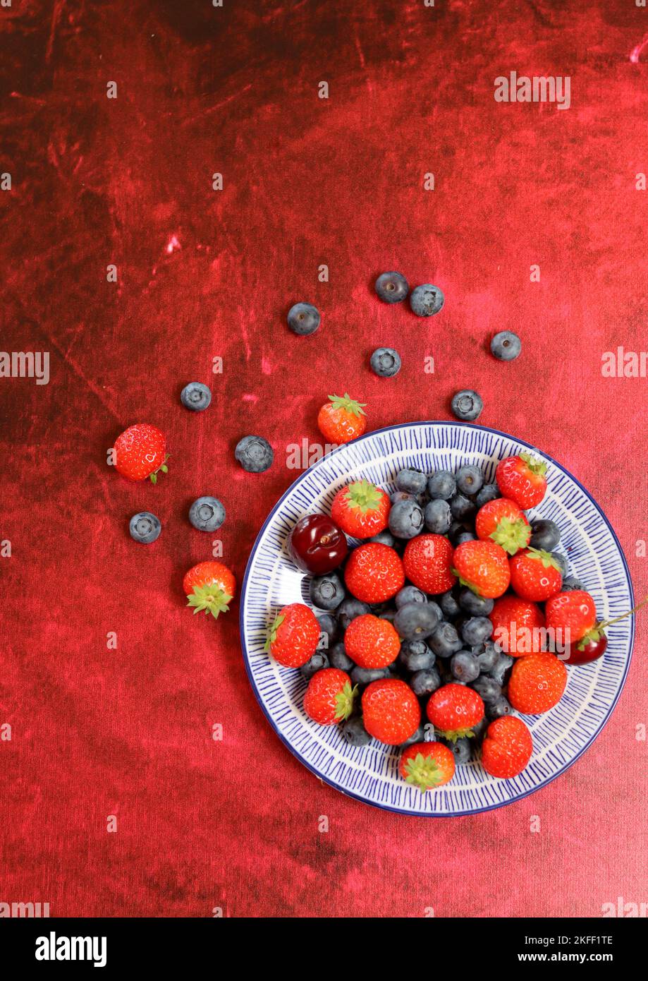 Vibrant colors of fresh summer berries. Strawberry, blueberry and sweet cherry on a blue ceramic plate. Red textured background with copy space. Stock Photo