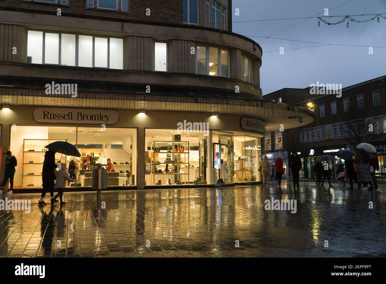 Exeter, UK, 16 November 2022: at dusk shop windows cast bright reflections across the pavements in Exeter as a band of heavy rainfall crossed the UK. Many shops are offering discounts for Black Friday and retailers are concerned that high inflation, the recession and the cost of living crisis will created a difficult trading environment for the retail sector. Anna Watson/Alamy Live News Stock Photo